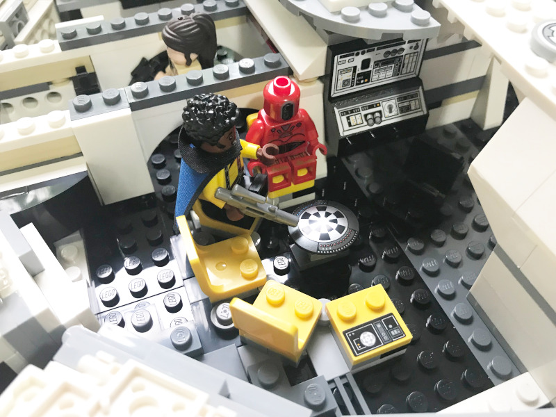 Detail from inside the LEGO Millenium Falcon. A Lego Lando is seated in a yellow chair in front of a round checkerboard table. A red droid is seated next to him. Behind a partition you can see the head of a woman.