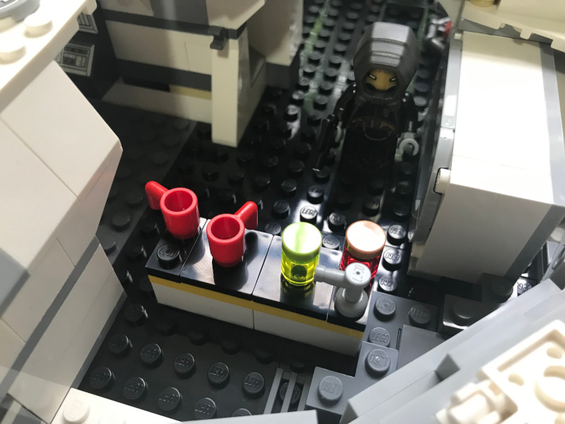 A LEGO figure cloaked in black standing in front of a LEGO countertop with two red LEGO mugs and two LEGO jars.