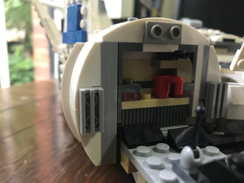 The cockpit without figures in it so you can see the back of the cockpit. There is a small shelf with a red mug on it and a kind of curved beige Lego piece of that with a black handle on top.
