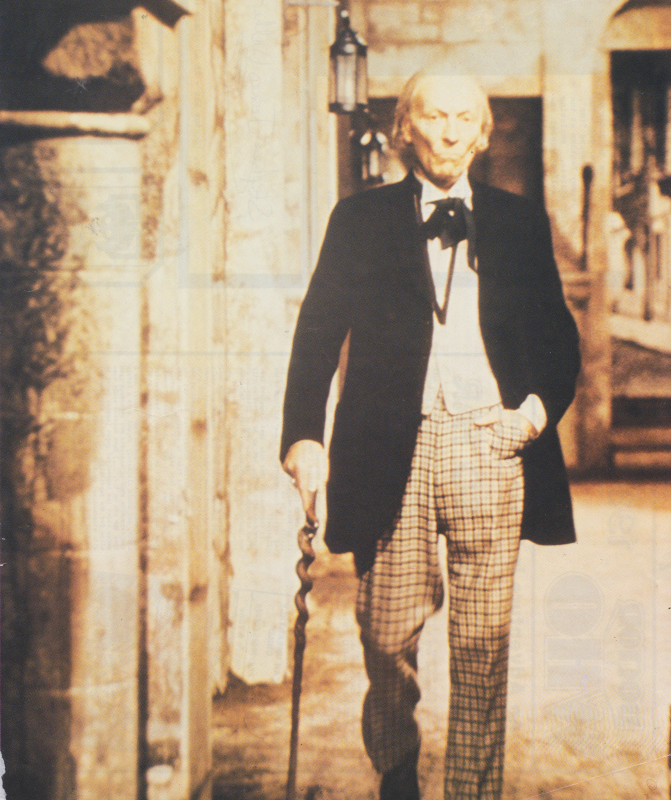 A rather sepia hued photo of the first Doctor walking down a street with a stick.