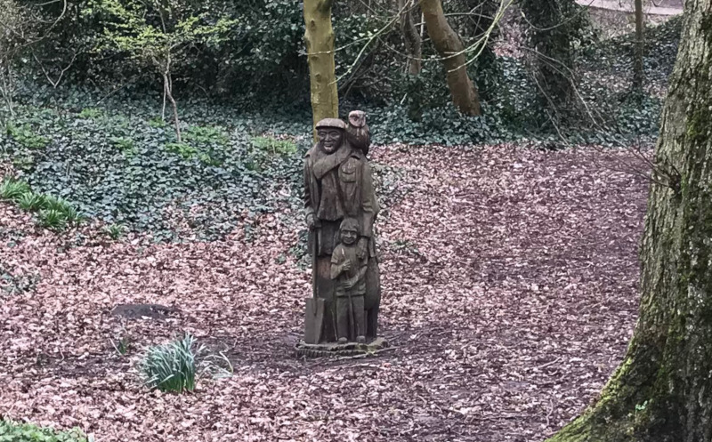 A wooden statue of a man holding a spade with an owl on his shoulder.  A small boy holding a stick stands in front of him.  The statue is in a wooded park area.