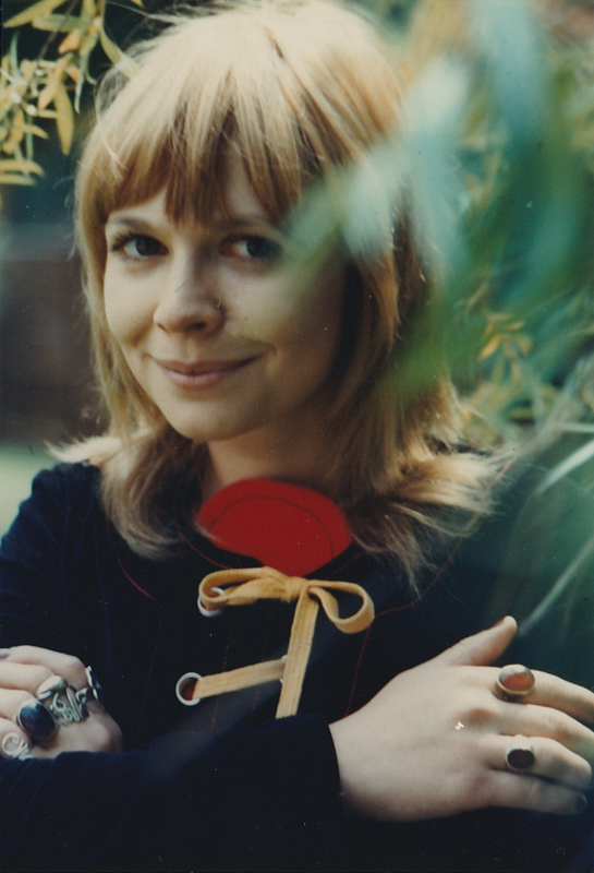 Head and shoulders publicity shot of Jo Grant.  She is wearing a black top with red and yellow detailing and has her arms folded showing lots of rings on her fingers.