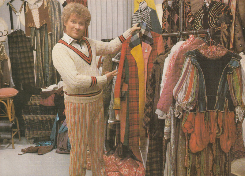 The Sixth Doctor, still in the cricket whites, is pulling his coat of the Tardis clothes rack and pointing at it.