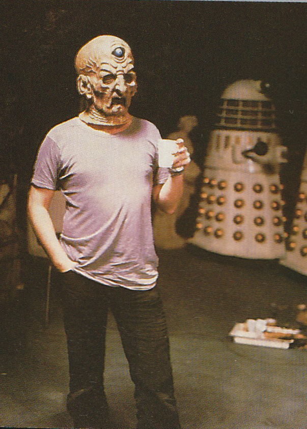 Man wearing Davros mask, a pink T-shirt and black jeans, drinking from a plastic cup with one hand in his jeans' pocket.