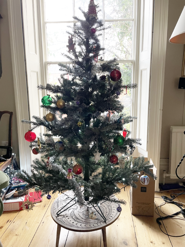 A decorated Christmas tree on a small table in front of a window.  A Dalek is in the fairy position.