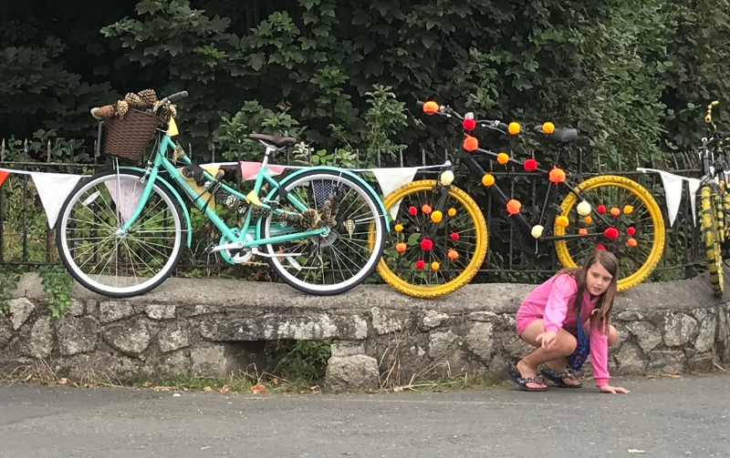 Two bicycles propped against railings on top of a low stone wall. One is blue with fir cones in the basket and one has yellow wheels and is decorated with red, orange and yellow pom-poms. A young girl in pink is crouched in front of them pointing at something.