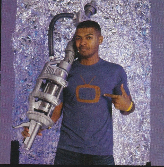Noel Clarke as Mickey from Doctor Who.  Publicity Still in front of a silvery background.  He's wearing a purple T-Shirt and carrying a big SF gun.