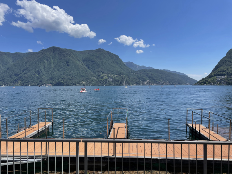 A picturesque lake in the sunshine with wooded mountains beyond.