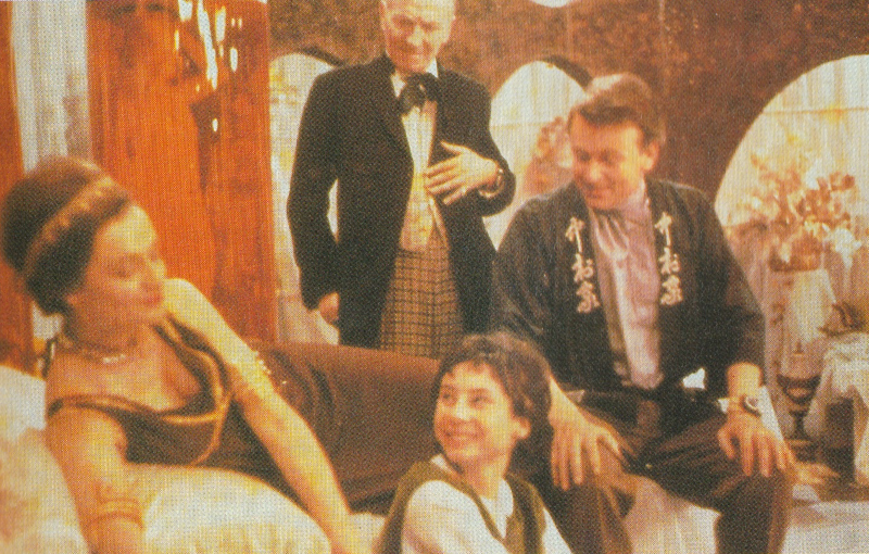 Slightly blurred colour photo from the Doctor Who episode The Keys of Marinus.  Barbara in a gown reclines on a couch.  Susan smiles at her while seated on the floor.  Ian, in a Chinese top is salt on the end of the couch also smiling.  The first Doctor gazes on benignly.