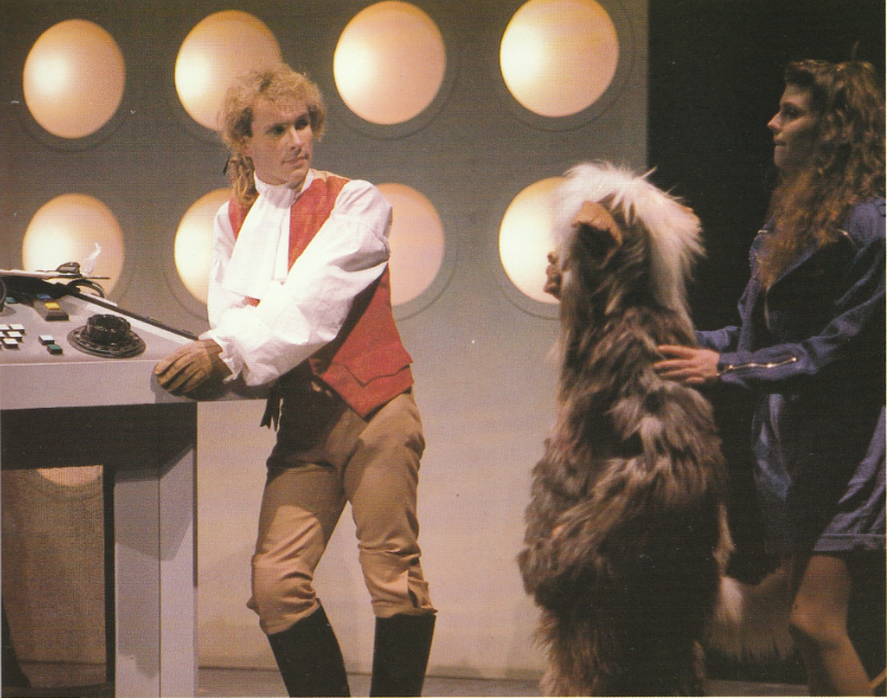 Interior of the TARDIS.  A man in French Revolution style clothing is at the controls.  A girl in a denim jacket and shorts looks on, her hands on the shoulders of a small furry creature - also looking on.