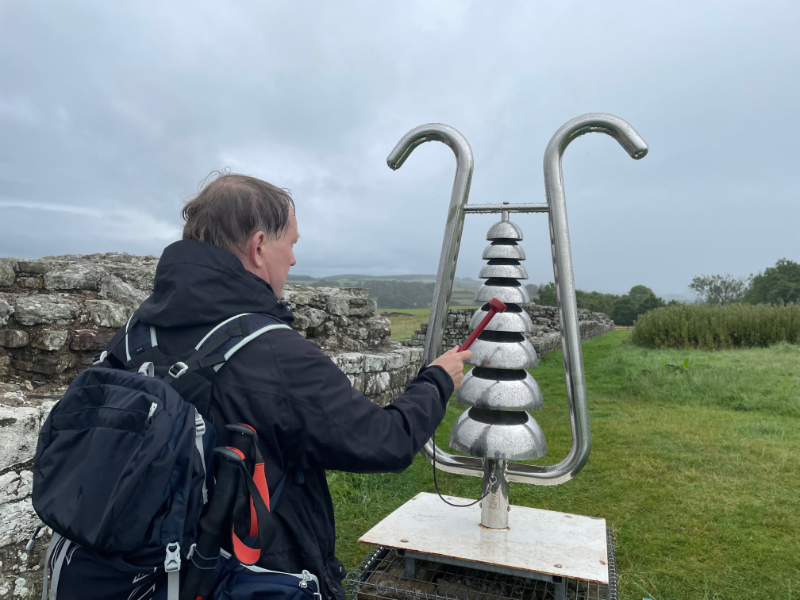 B standing next to some wall.  He is hitting a large silver thing that looks a bit like a giant metal lyre with curved downward facing bowls where the strings would be.  He is hitting it with a small red hammer attached to it with string.