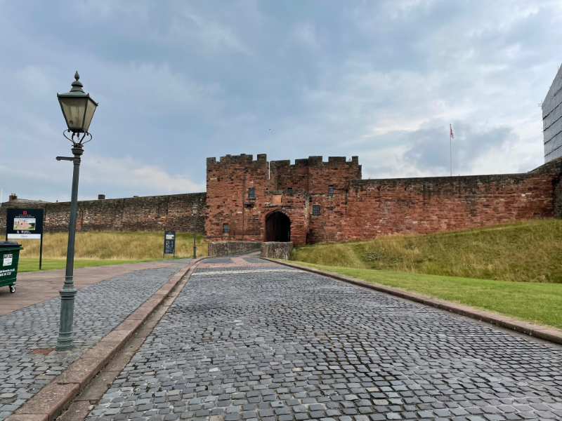 Red brick walls with a red brick gateway with battlements.  A cobbled walkway with a modern looks leads up to it.
