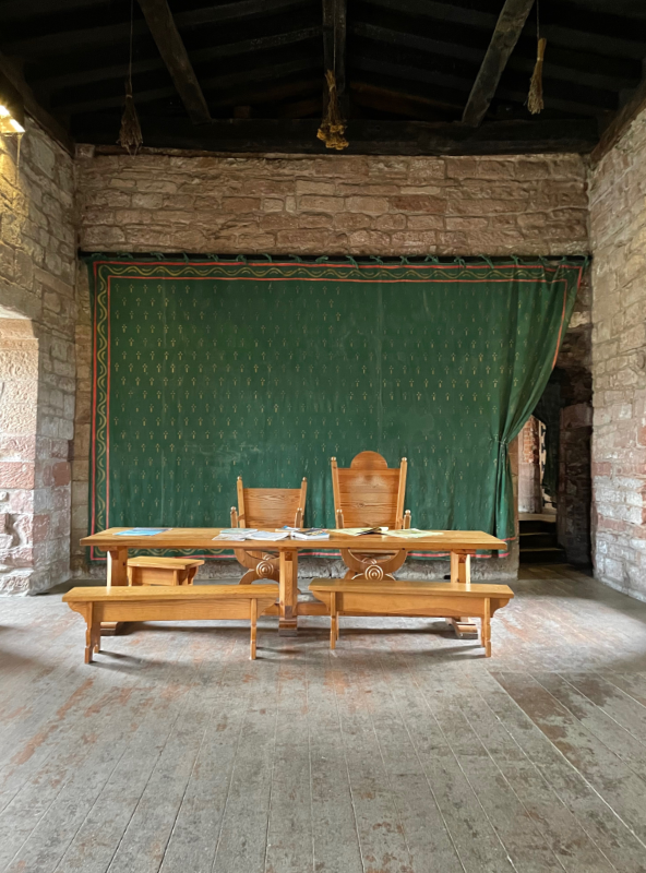 A stone room with a wooden floor. In front of a green curtain that dominates one wall is a long wooden table. Two benches are this side of the table and two carved wooden chairs the other.
