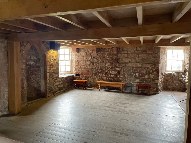 A stone room with a wooden floor. Wooden eaves look new. A small bench and table stand at the far side, next to a glass paned window.