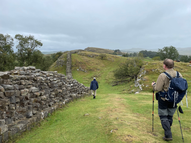 Back view of B and another walker on rolling hills with the remains of Hadrian's wall to the left.