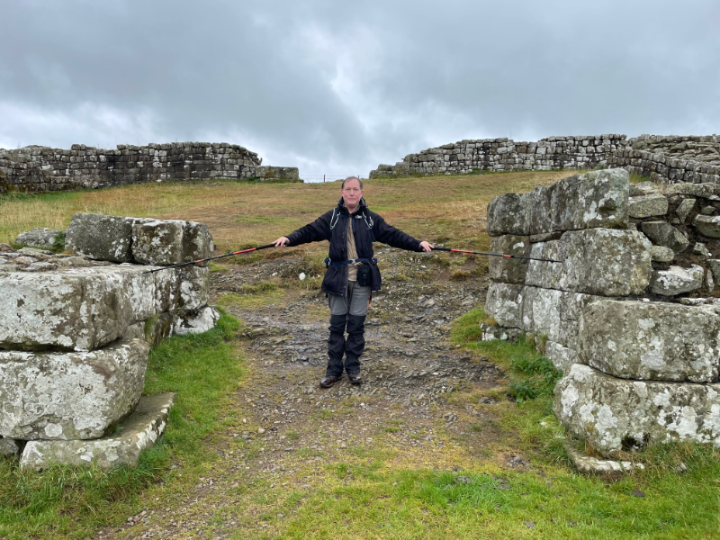 B stands between two stone walls, touching each one with the tips of his walking sticks. It is clearly a square structure and the four sides can be seen behind him.