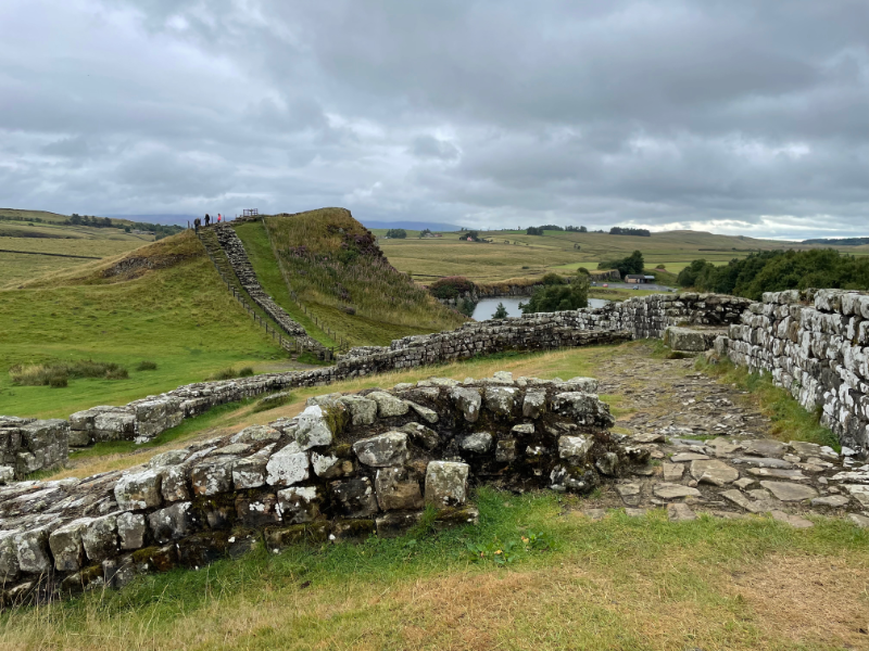 A view of a square milecastle close too with the wall stretching beyond down into a dip and then up again.
