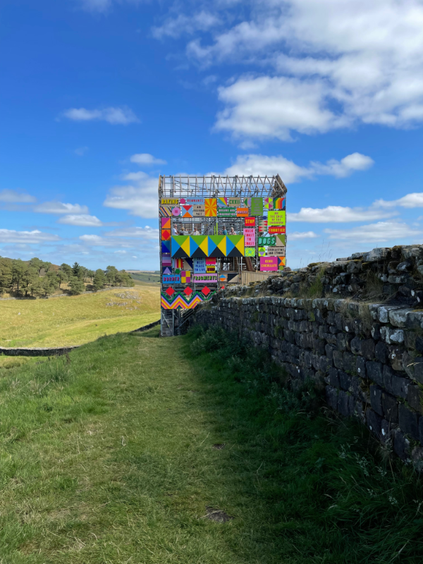 The gatehouse from the side as it straddles the wall of the fort.  Scaffolding is visible with a sloping roof with no covering on the framework.  Colourful boards in pinks, greens, reds and blues are visible.  Particularly dominant is a rectangular board with a pattern made up of black, yellow, blue and green triangles.  Some of the panes have text on them but the resolution isn't really good enough to read them.  On says STOP in yellow on a red background.  Another says DOGS in blue on a green background.