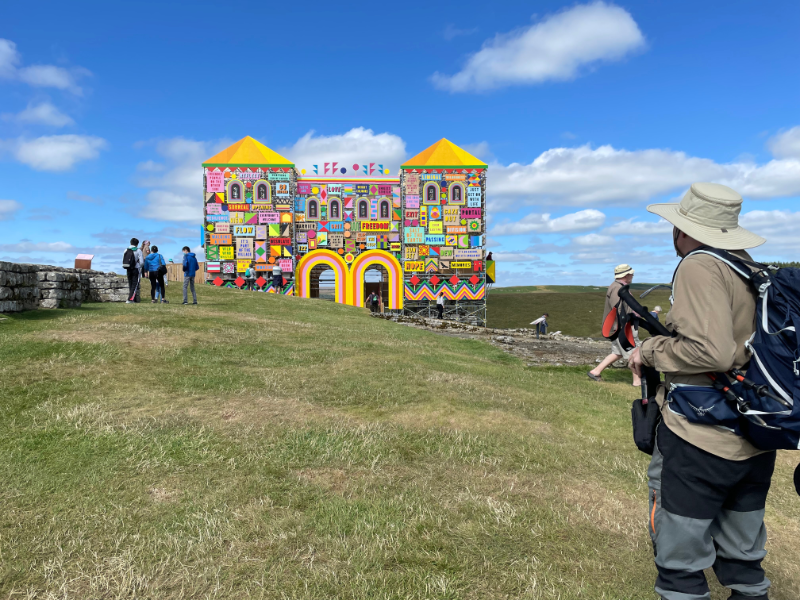 B admires the gatehouse from within the fort.  It has two turrets either side of the main entrance which consists of two arches in yellow, pink and red stripes.  Small arched windows are visible on the second storey and the pointed turret roofs are in yellow and orange.  More colourful boards decorate the sides.  Most of the signs are too small to read, but one says Freedom in red on a yellow background.