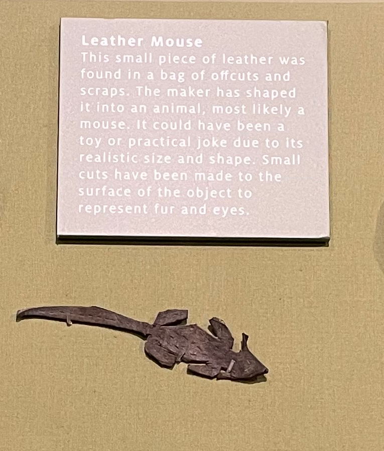 Small leader mouse in a display case.  The legend reads: Leather Mouse.  This small piece of leather was found in a bag of offcuts and scraps.  The maker has shaped it into an animal, most likely a mouse.  It could have been a toy or practical joke due to its realistic size and shape.  Small cuts have been made to the surface of the object to represent fur and eyes.