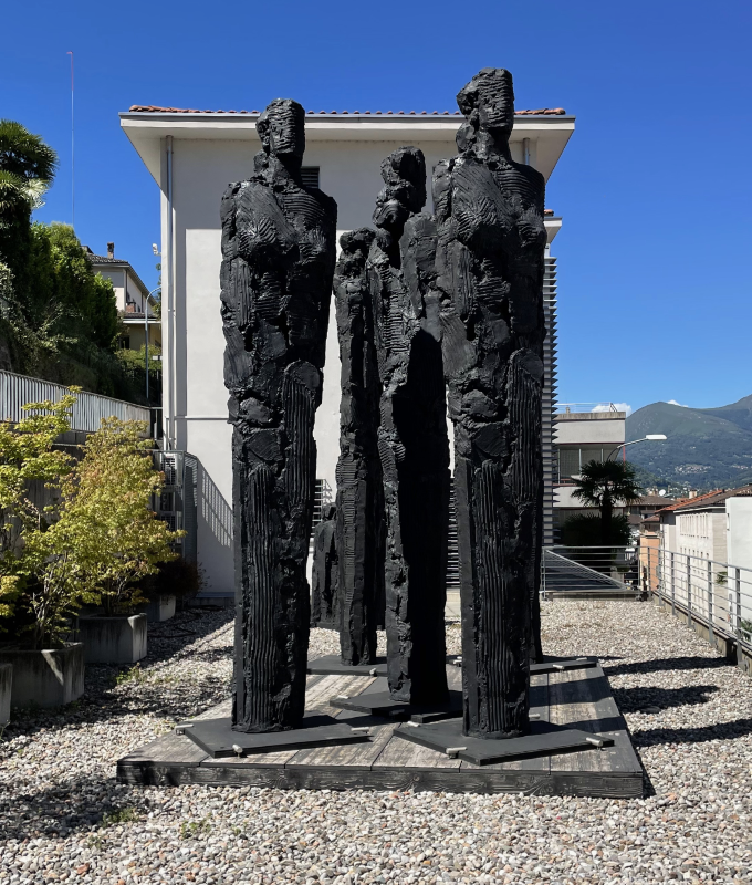 Four tall black vaguely human sculptures surrounded by gravel.