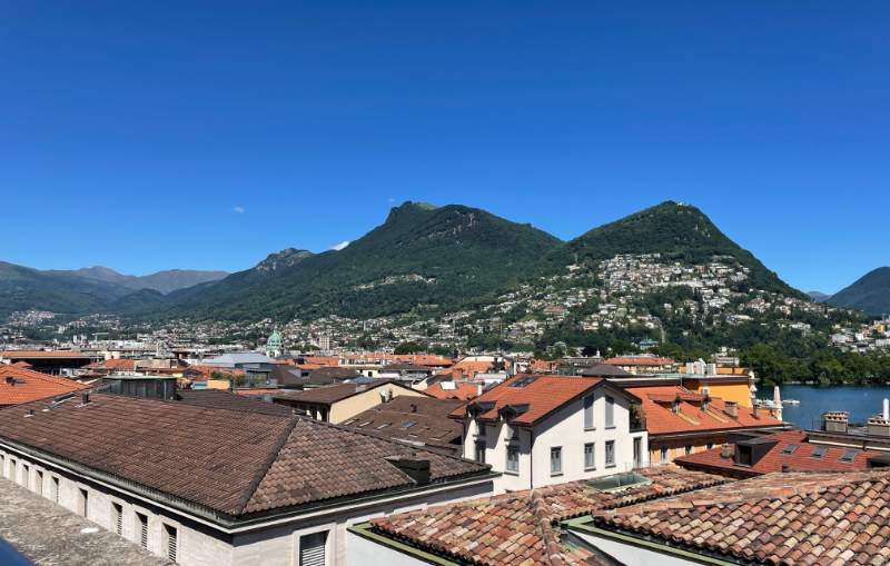 View over tiled rooftops, to a blue lake with wooded mountains beyond.