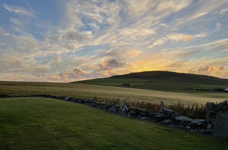 A evening sky with yellowish clouds behind a low hill and dry stone wall.