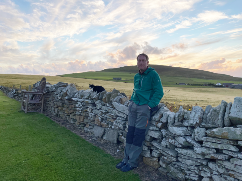 B. leaning against a dry stone wall with a cat on it. A hill is in the background and wooden chair is placed against the wall.