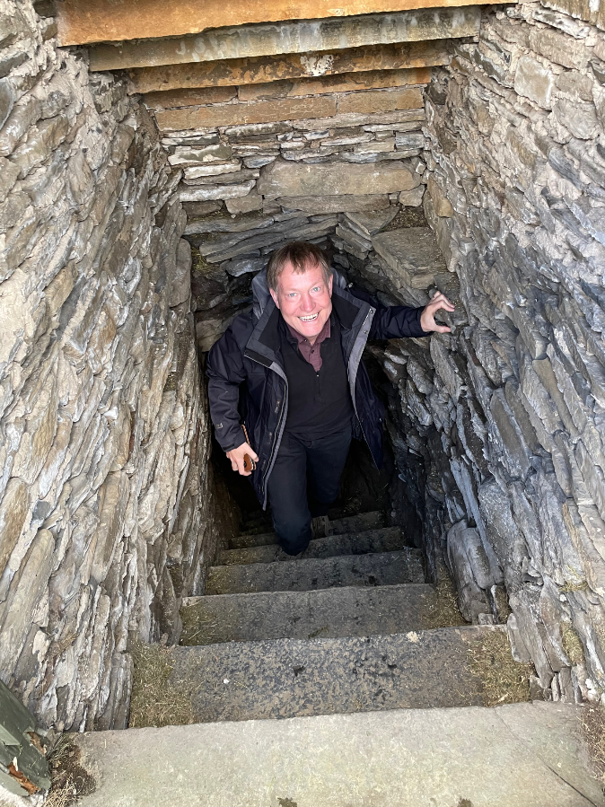 B emerging with a grin up concrete steps but with stones lined walls and ceiling.