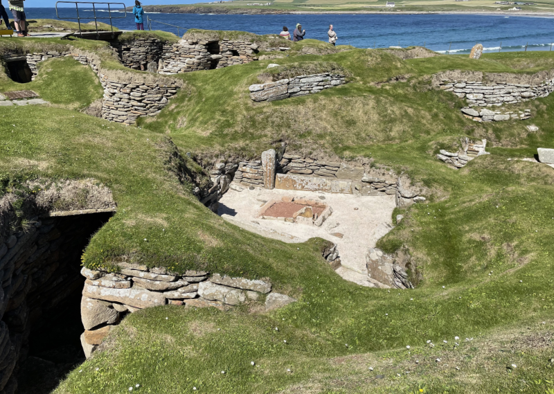 Grass covered sand dunes, revealing walls and sandy floors, in the centre a room with a hearth.