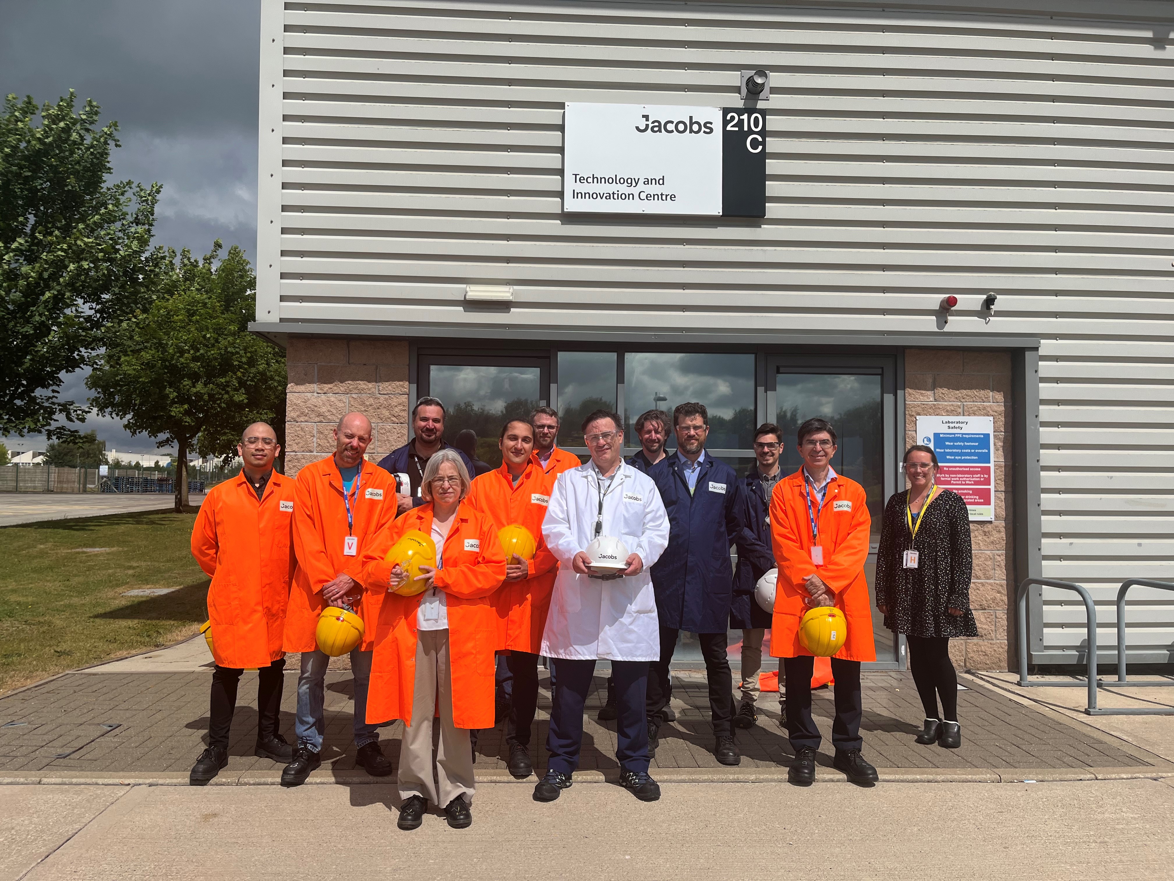 A group of people in overalls holding hard hats, squinting into the sun.  Most of the overalls are bright orange.  I am at the front.  We are standing in front of a building signed Jacobs 210 C Technology and Innovation Centre.