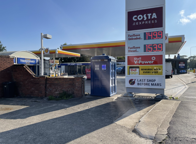 A Tardis next to a sign advertising petrol costs and a costa shop.  A Shell garage is in the background.
