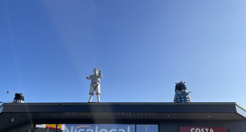 A Cyberman stands, on arm upraised, on a flat roof, flanked by Daleks.
