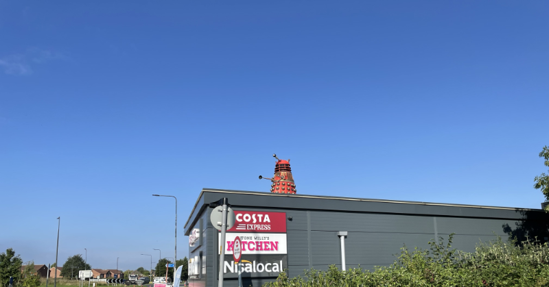 A red dalek stands on a flat roof.  Below it a sign advertises Costa, a Kitchen shop and a Nisa Local.