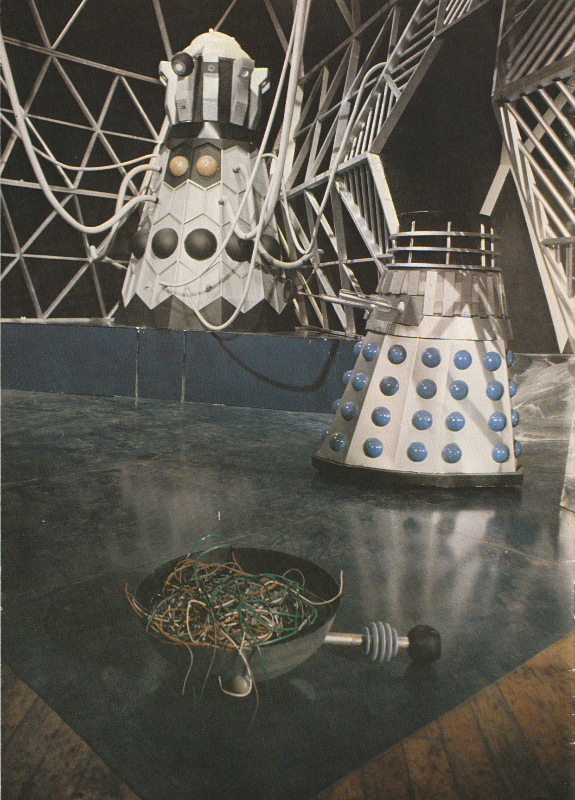 The Emperor Dalek from Evil of The Daleks on his podium. A Dalek in the foreground with the top of its head removed. This is upside down on the floor containing what looks like a bunch of wires.
