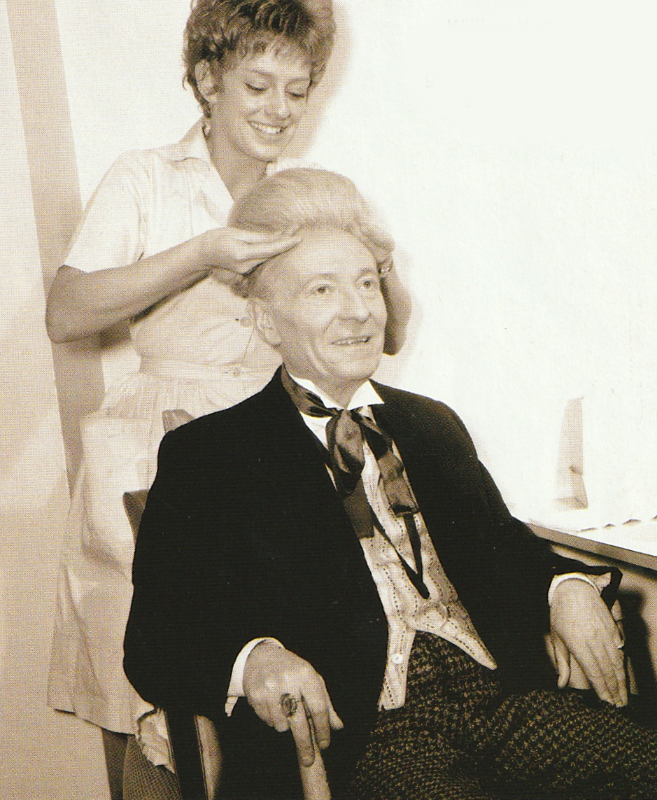 William Hartnell sitting in a make-up chair while a lady places his Doctor Who wig on his head.