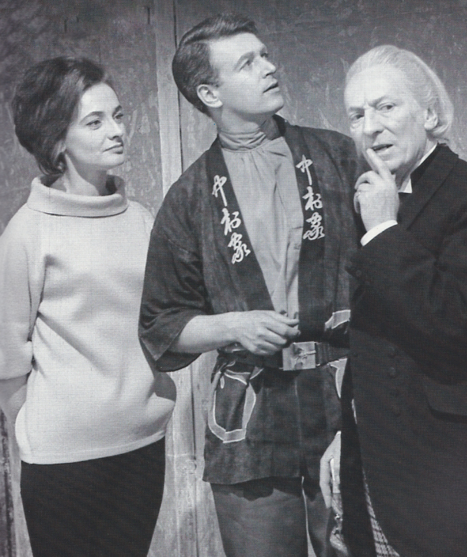 The first Doctor, Ian and Barbara in the Keys of Marinus.  The Doctor looks to camera with one finger held up to his mouth as if to shush someone.  Ian wears a loose jacket, belted at the waist, with Chinese characters decorating the edges.  He is looking up at something past the Doctor.  Barbara is wearing a wide-necked polo neck top.  She is looking at the Doctor.