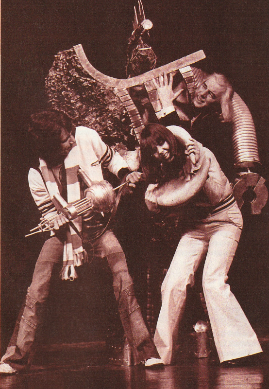 Black and white photo, sepia tinged.  Some kind of large monster grips two people.  In the back a man with a longish white wig some kind of metal claw hangs down behind him.  In the front Wendy Padbury has an organic-looking claw around her neck and is wearing flared trousers.  Next to Wendy a young man with a short scarf around his next is holding some kind of device (or possibly another of the monsters appendages).