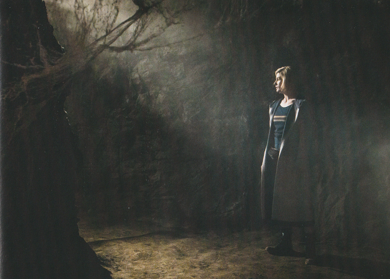 The Thirteenth Doctor in a cave with light coming in from above.  Cobwebs in the foreground to the left of the image make it look a little like there is a tree there.