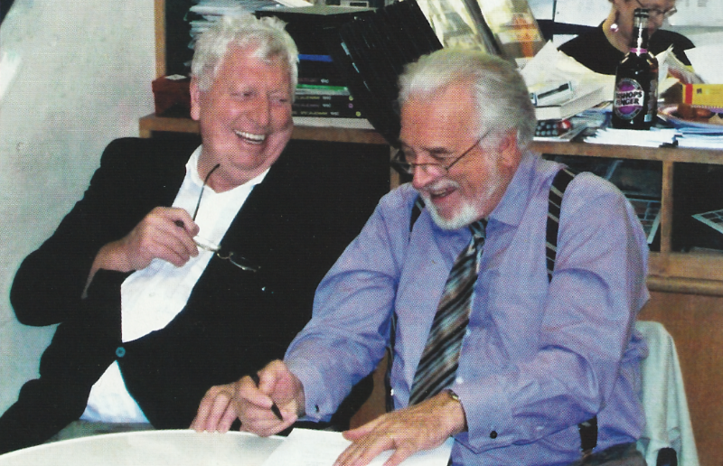 Tom Baker and Nicholas Courtney sitting side by side behind a table probably some time in the early 2000s - both are white haired, wearing (or in Baker's case holding) glasses.  Both are laughing.  Courtney is holding a pen and appears to be signing something.  Tom baker is looking at Courtney.