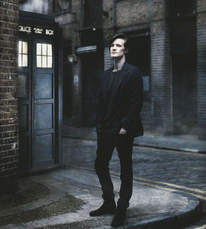 Publicity shot of Matt Smith in a dark jacket and trousers, in a dimly lit alley with the Tardis in the background.