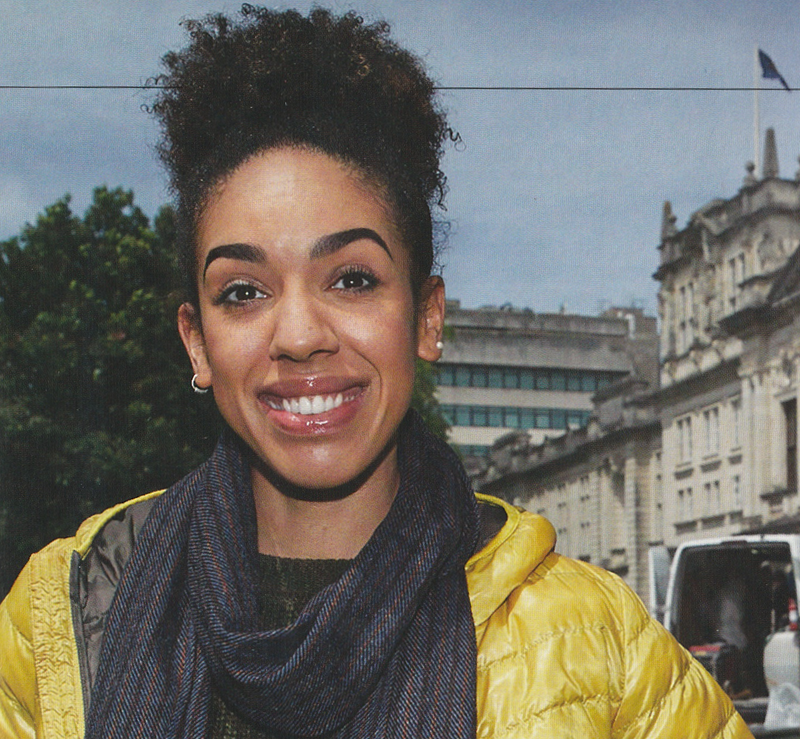 Photo of Pearl Mackie behind the scenes in Doctor Who.  She is wearing a yellow puffer jacket with a grey scarf around her neck and has her hair up.