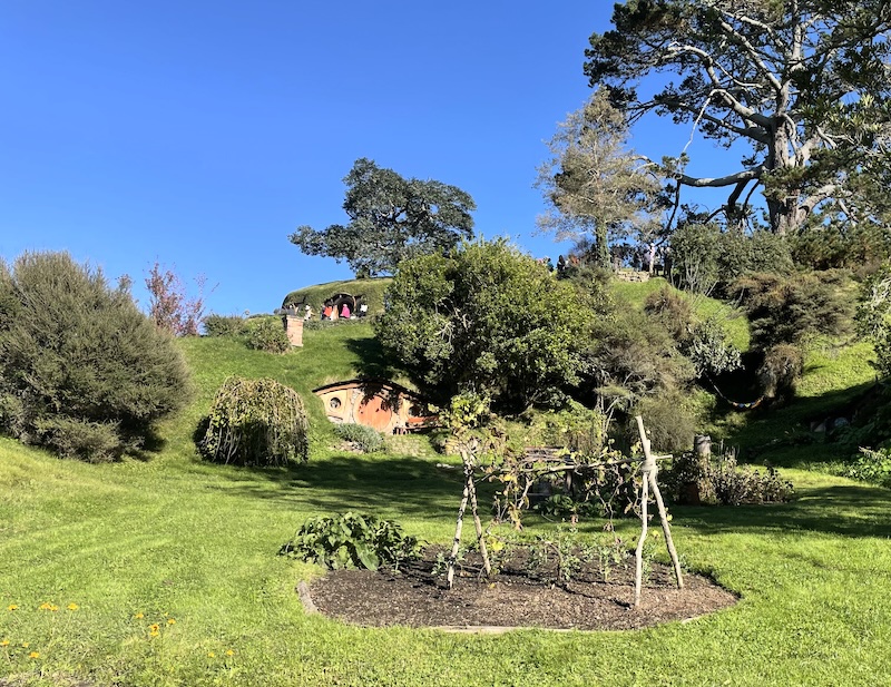 Some kind of frame for climbing plants in front of a a hobbit hole set into a hill with a second hobbit hole visible higher up the hill.