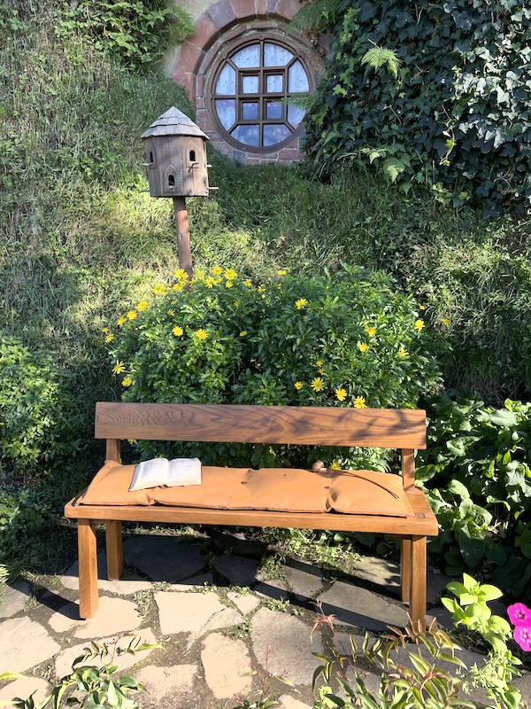 A bench with a book and a pipe on it, some kind of bird house behind and a window in the hill beyond that.