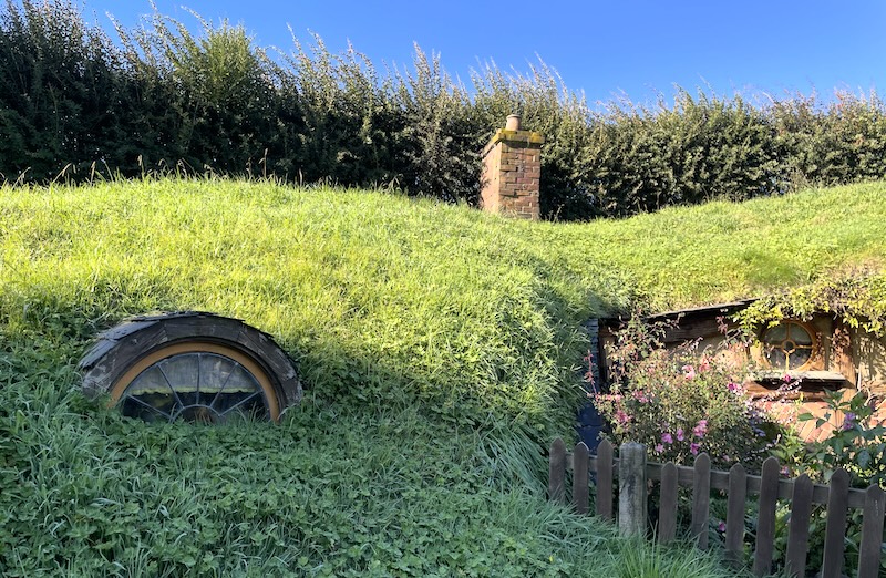 An arched window peeping out of the grass, next to a fenced in garden and a chimney on top of the rise.