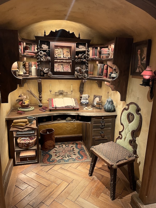 A nook containing a writing desk and chair. Above the desk are shelves crammed with books and scrolls.