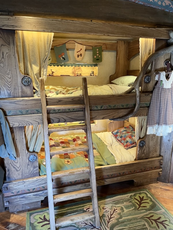 Wooden bunk beds with a ladder. Colourful patchwork quilts and pillow. A dress hangs up to one side.