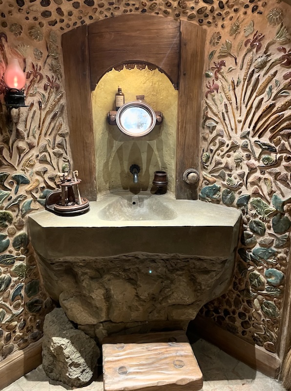 A stone wash basin with a little circular mirror mounted in an alcove above it. Decorative plasterwork either side shows pond leaves a frog and rushes.