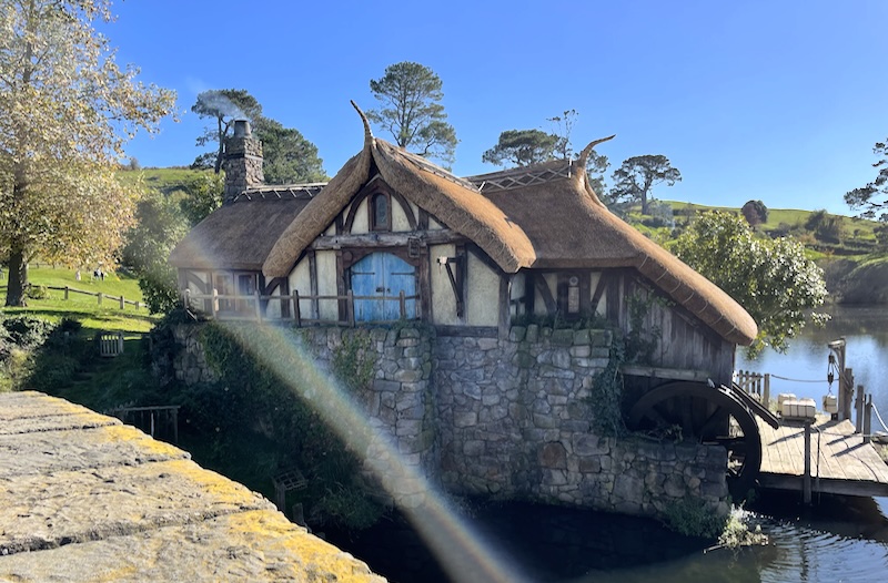 A thatched cottage raised on a stone base above the lake. A mill wheel is on one side below the cottage next to a jetty.