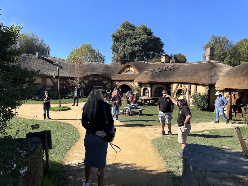 A courtyard full of tourists (and guides). A long low thatched building with arched doors and round windows beyond.